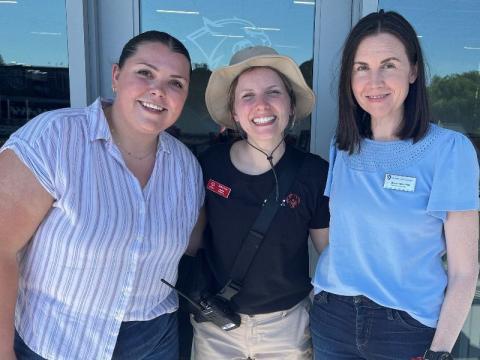 Three women standing outside on a sunny day, smiling and posing together for a picture.