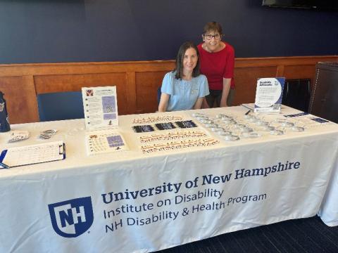 Two women behind a table with a tablecloth that reads "University of New Hampshire Institute on Disability NH Disability and Health Program".