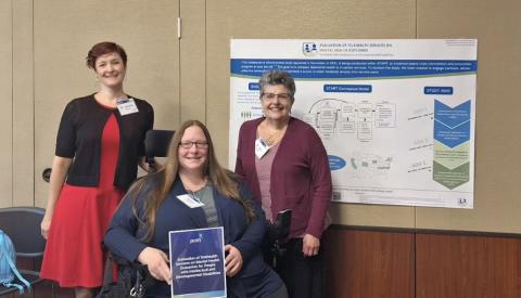 Three people, including one person in a wheelchair, in front of a poster they authored about the telehealth study