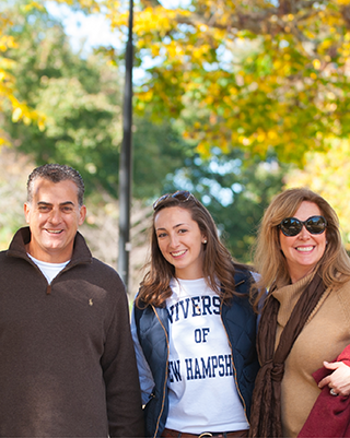 a smiling mom, dad, and daughter wearing a UNH tee shirt outside on a sunny fall day