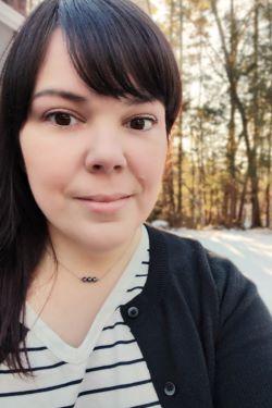 Rita Despres is a woman in her mid-forties is outdoors, looking at the camera. She has dark hair, dark eyes, and is wearing a black and white striped shirt with a black cardigan over it. It is winter, and there is snow on the ground. The sun is starting to rise between the trees behind her.