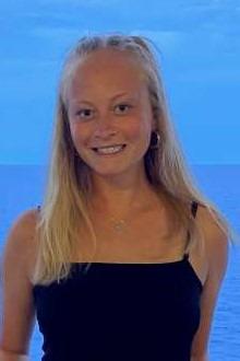 Kathryn Hughes is a woman with straight blonde hair wearing a black dress, pictured outside in front of the ocean.
