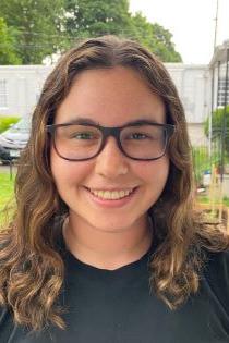 Caroline Lambert is a woman with brown hair wearing glasses, pictured outside in front of a white building surrounded by trees.