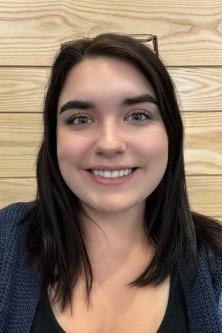 Alyssa Limeburner is a woman with dark hair and green eyes. She is wearing glasses on top of her head, a black shirt and a blue toned cardigan. She is pictured in front of a light wooden background.
