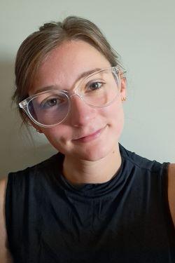 Abigail Morrow is a woman with brown straight hair in a bun, wearing clear glasses, a black shirt, pictured in front of a light-yellow wall.