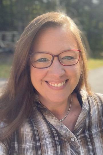 Karin Mortimer is a woman with light brown hair, and she is wearing red framed glasses. She is pictured standing in her yard with her driveway and trees behind her. The sun is shining on her, and she is smiling.