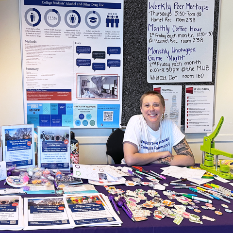 Heather Morris, a young white person with a buzzcut and a white t-shirt, smiles while hosting a resource table arrayed with flyers, candy, buttons, and stickers