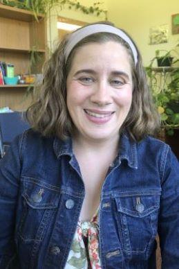 Lara Raymond is a woman with wavy brown hair wearing a jean jacket, pictured in an office in front of many climbing plants.