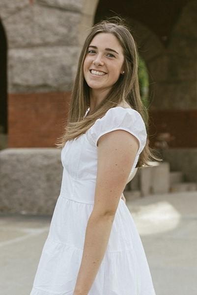 Lauren Reid is a woman with light brown hair wearing a white dress pictured in front of UNH T-Hall.