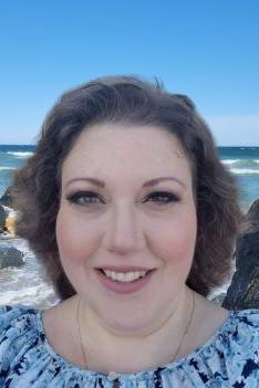 Nicole Sheaff is a Caucasian woman with brown wavy hair, wearing a blue and white printed shirt, pictured in front of a rocky beach and ocean waves.