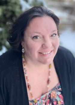 Lisa Steadman is a white woman with brown hair in her 40s in a dark floral top in front of a pine tree.
