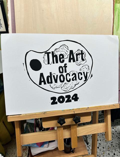 A wooden easel holds a white artboard with an black and white image of a paint palette and text reading The Art of Advocacy 2024