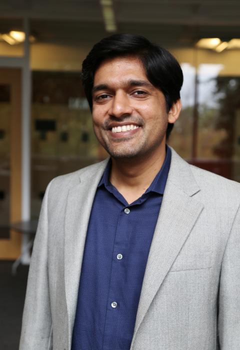 Kartik Trivedi is a South Asian man with straight black hair with grey streaks, here standing indoor in front of a glass wall. 