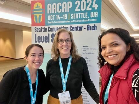 Three women in professional attire standing in front of a sign at a conference and smiling for a group photo