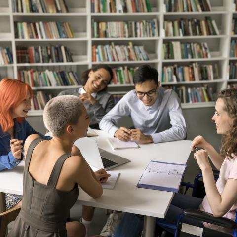 A group of five diverse young adults gathered around a table in a library or study space, smiling and engaged in discussion. They are surrounded by shelves filled with books. One person in a wheelchair is part of the group, contributing to an inclusive and collaborative atmosphere.