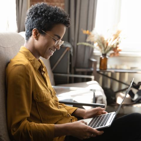 A young woman with short curly hair and glasses sits comfortably on a couch, smiling as she works on a laptop. She is wearing a mustard yellow shirt and appears relaxed in a cozy, well-lit room with soft natural light.