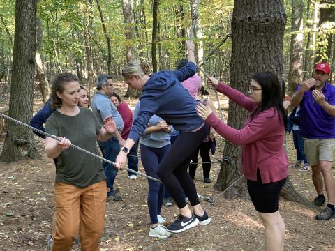 Two people help a third woman balance on a tightrope while she makes her way across. In the background, another group is doing a similar activity.