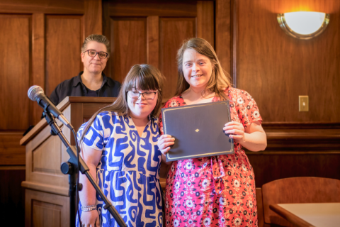 Listening to shared experiences: Two young women with disabilities stand smiling together next to a microphone in a courtroom, while an older woman from behind a podium looks at them and smiles.