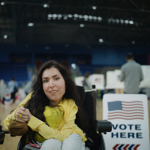 A woman in a power wheel chair poses next to a Vote Here sign with the American Flag at a polling center.