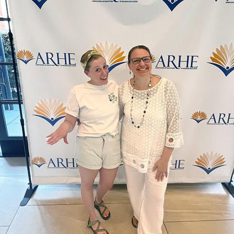 Heather Morris and Heidi Cloutier smile for the camera in front of an ARHE step-and-repeat backdrop at the ARHE Conference.