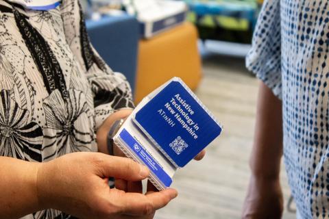Someone holds a cellphone or tablet holder made out of corrugated plastic. It's branded with information on the Assistive Technology in New Hampshire (ATinNH) program.