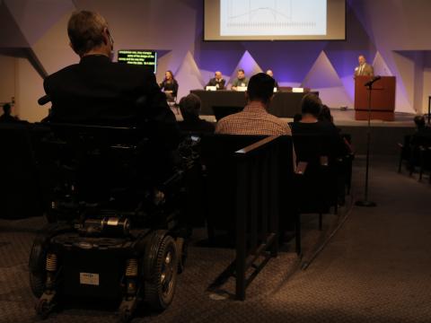 A person in a power wheelchair is part of an audience watching a presentation on a stage. The presentation has powerpoint slides, CART, and ASL interpretation and the speaker has is using a mic. All of these are types of assistive technology in use.