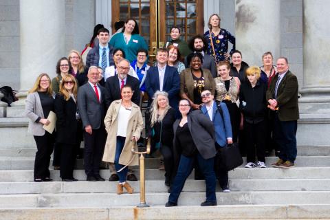 A group of diverse people stand on the steps to the NH State house smiling and posing for a group photo.