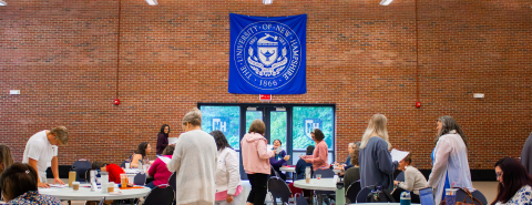 A large group of people are working and collaborating in one of the UNH event spaces, while a large blue banner hangs over the door with the words "The University of New Hampshire -1866 - 1893 1923 Science, Arts, Industry" with accompanying symbolism