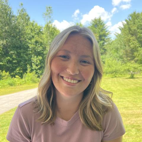 Alyssa Gagne is a woman with medium length blonde hair pictured outside in front of some trees. 