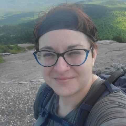 Zee Immele is a person with short brown hair wearing glasses. She is standing on top of a mountain with hills in the background.  