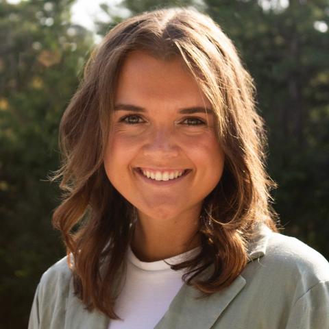 Maggie Mahoney is a woman with brown hair and hazel green eyes standing outside in front of some trees 