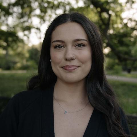 Sarah Perry is a woman with brown wavy hair, pictured outside in front of some trees. 