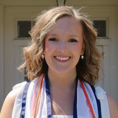 Audrey Young is a woman with brown hair wearing a white dress, pictured outside in front of an academic building. 