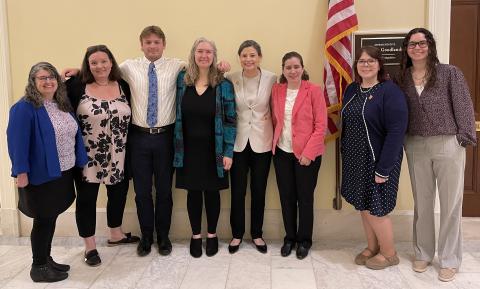 A group of eight professionally dressed people smile together in front of the sign to NH Representative Maggie Goodlander's DC office placard