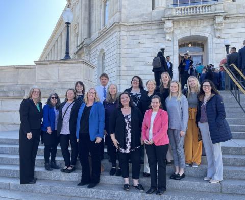 A group of happy professionally dressed people stand outside together on the steps to the US Capitol Building.