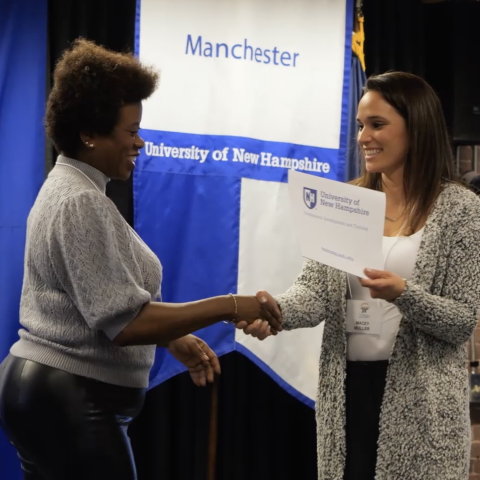 Two women in front of a banner that says "University of New Hampshire - Manchester" shake hands as one gives the other a certificate.