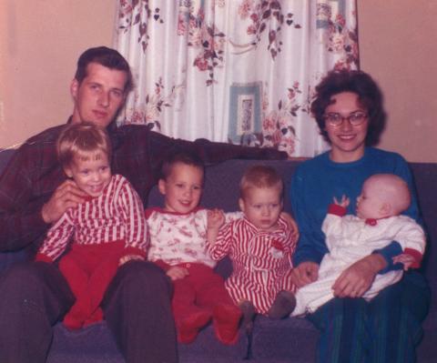 Two young parents pose on a couch indoors with their four children who look to all be under the age of 5. The children are all wearing outfits with red and white, potentially for the holidays.