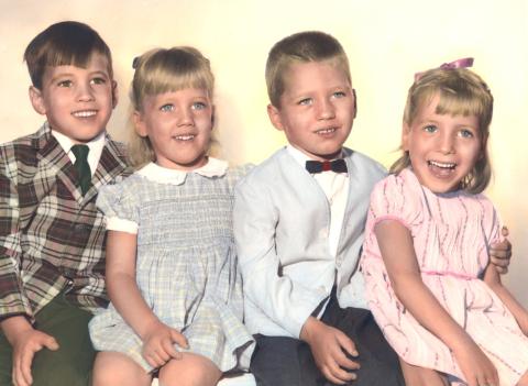 Four young siblings, two girls, two boys, pose for a family photo. They appear to be between the ages of 4 to 9. The boys are wearing nice jackets and ties, the girls are wearing dresses.