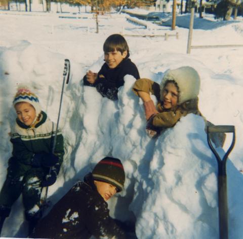 Four young kids pose together on a snow fort they built that is about as tall as they are.