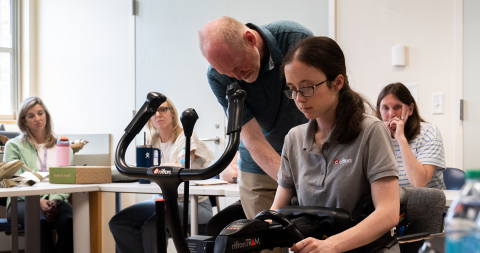 A man and woman demonstrate some assistive technology equipment to a room full of people.