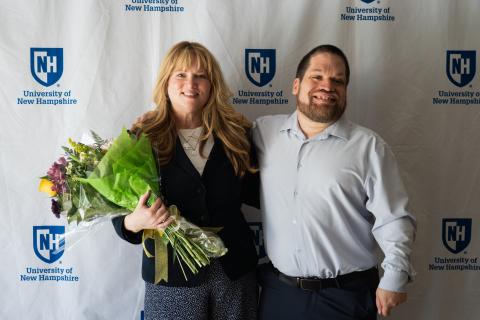 Tim Houle standing next to a lady, Tim has her arm around her and they are smiling for a photo. The lady next to Tim is holding a boquet of flowers. They are standing in front of a white backdrop with the University of New Hampshire logo on it. 