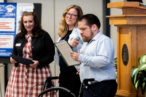 Tim Houle speaking into a microphone while reading from a piece of paper. Two Woman Stand next to him looking at him and smiling. 