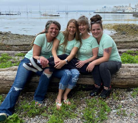 A group of four women in light green tee-shirts that say Reentry Sisters sitting on a log in front of a shoreline.