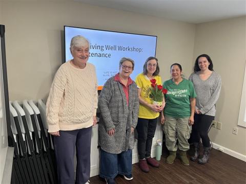 Five women of various ages stand together in front of a screen that says "Living Well Workshop: Maintenance"