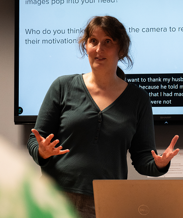 A woman at a podium presents to her audience, talking animatedly with her hands.