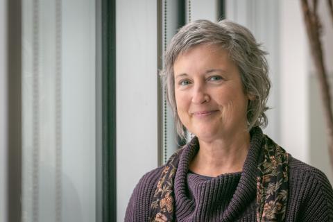 Tracey Miller is a woman with wavy midlength gray hair, wearing a purple sweater with a floral scarf, sitting in front of windows.