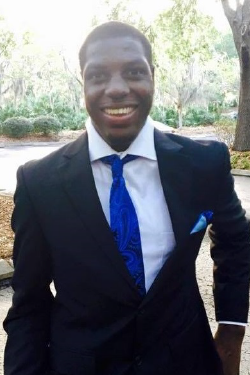 Headshot of Cyril Nonga-Mann smiling, with short dark hair, wearing a dark suit and white button-up shirt with a purple tie. 