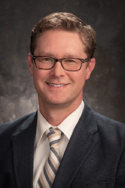 Headshot of Eric Moody smiling, with short brown hair, wearing glasses and a dark suit with white button up shirt and striped tie. 