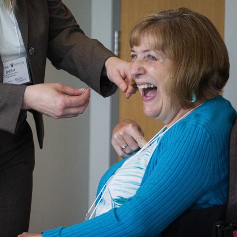 A woman with a blond bob wearing colorful clothes and seated in a wheelchair, laughs delightedly as someone goes to pin something to her shirt.