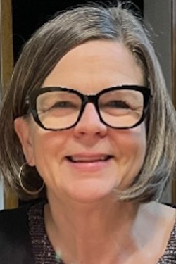 Headshot of Karla Ausderau smiling, with chin-length hair, wearing glasses and a black cardigan over a dark top. 